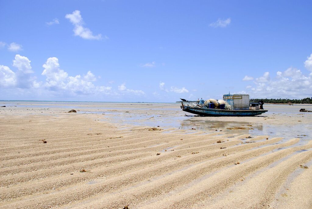 Vista paradisíaca da Praia do Patacho com coqueiros ao longe, areia branca e um barco na areia.