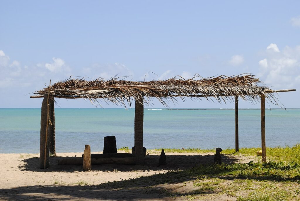 Vista de uma cabana de palha na areia da Praia de Patacho com o mar ao fundo.