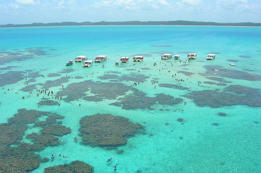 Vista aérea do mar de Antunes, com águas azuis e corais com turistas em barcos.